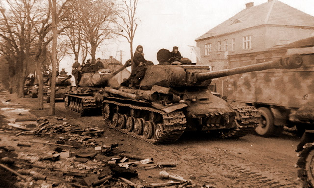 Колонна тяжёлых танков ИС-2 проходит через населённый пункт где-то в Восточной Пруссии. По обочине разбросаны больше не опасные танкистам фаустпатроны